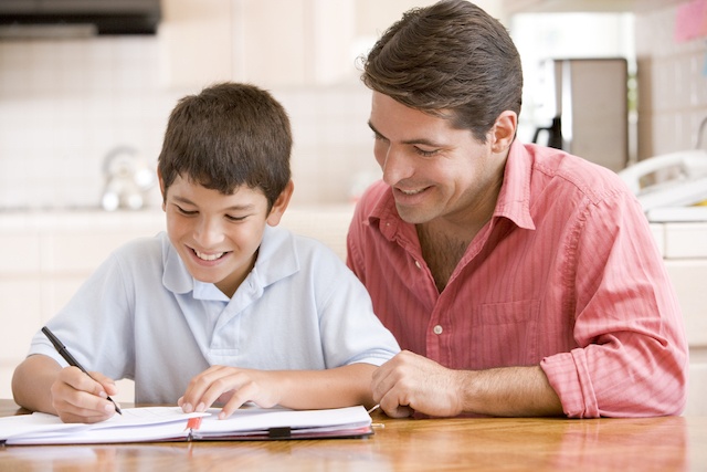 Parent guiding a smiling boy as he practices writing at the kitchen table